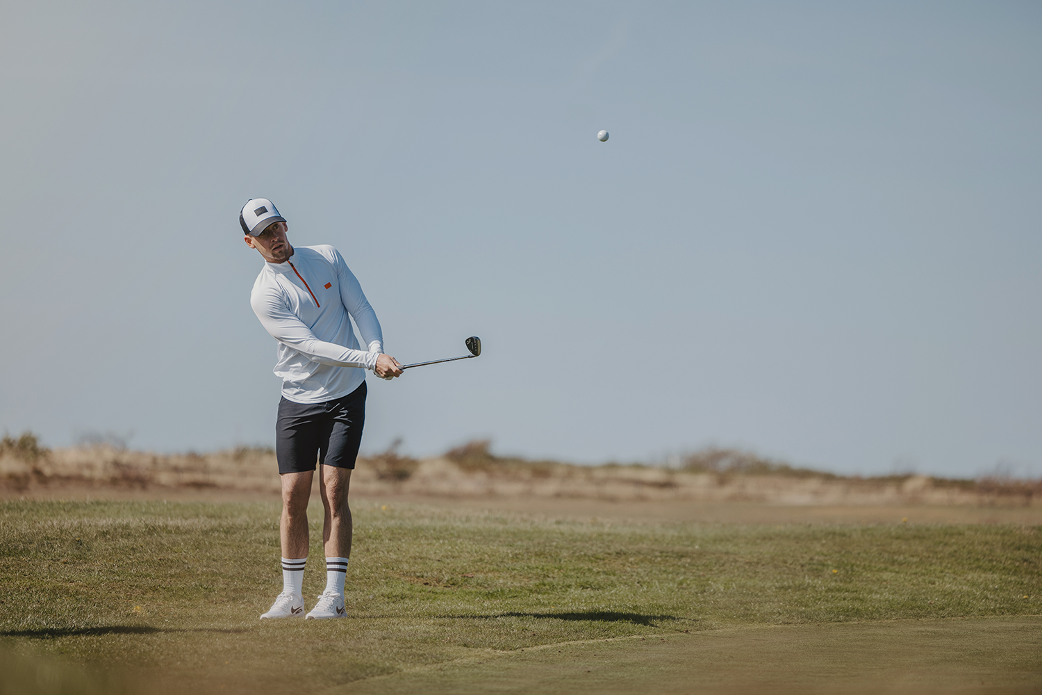 Man slår en golfbold med en kølle på en golfbane, iført en hvid trøje, shorts og kasket under en klar himmel.