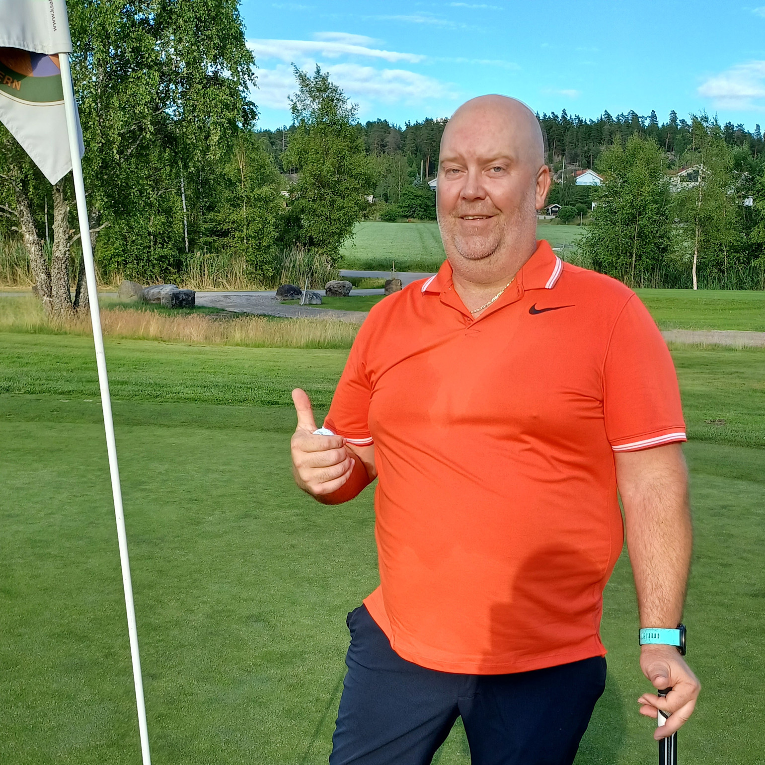 Mand i orange polo giver tommelfingeren op og holder en golfkølle på en golfbane med grønt og et flag i baggrunden.