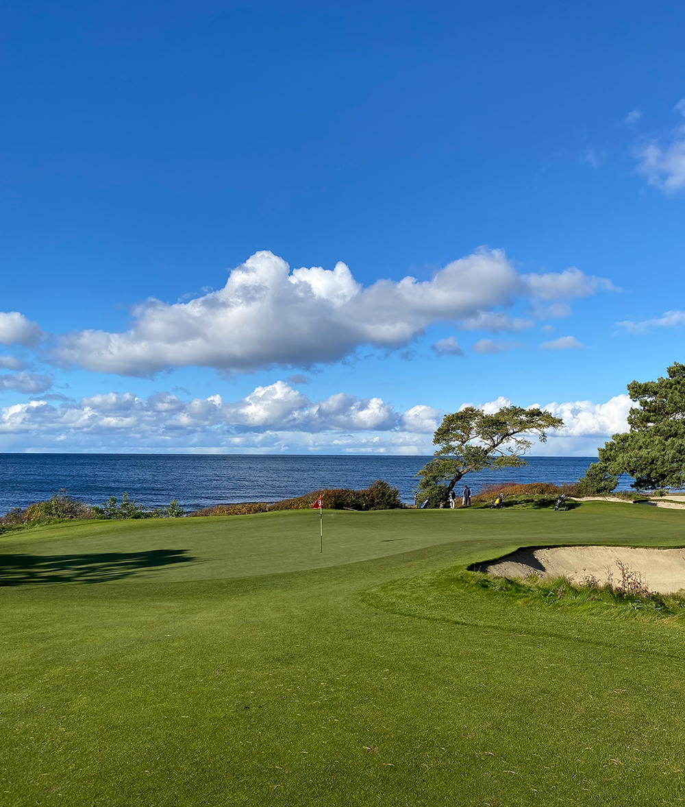 Golfgreen med flagga, sandbunker och hav i bakgrunden under blå himmel med vita moln.