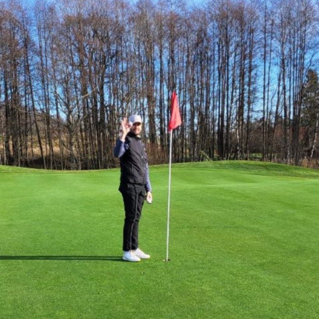 Person står bredvid en golfflagga på en grön golfbana, gör OK-tecken med handen, träd i bakgrunden.