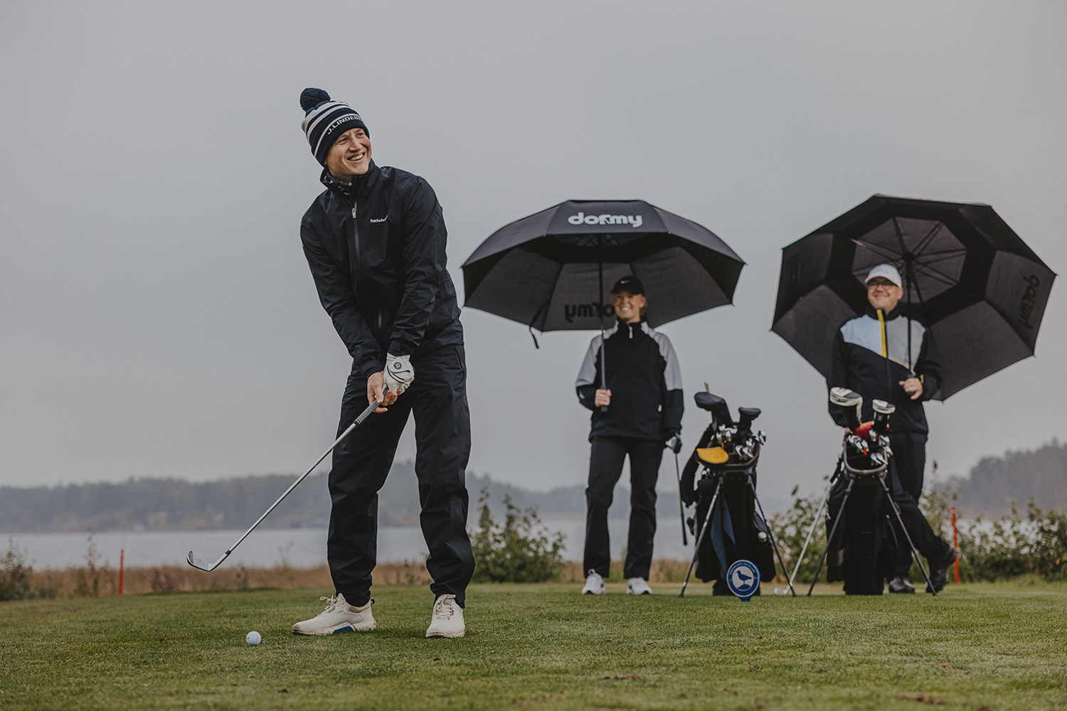 En golfspelare svingar klubban mot en boll medan två personer med paraplyer och golfbagar tittar på vid en gråmulen golfbana.