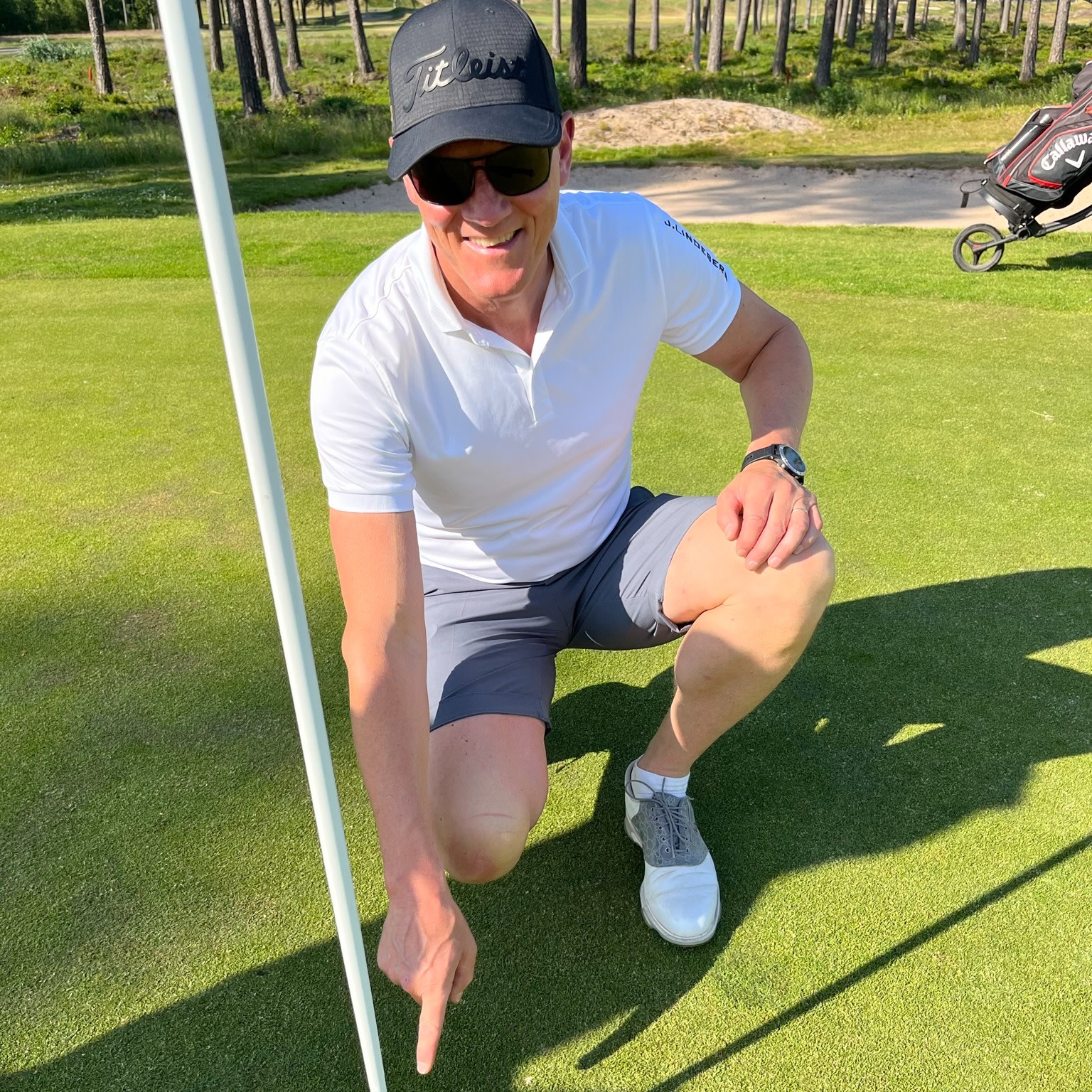 Un homme en polo blanc et casquette sourit et pointe du doigt le trou sur un green de golf par une journée ensoleillée.