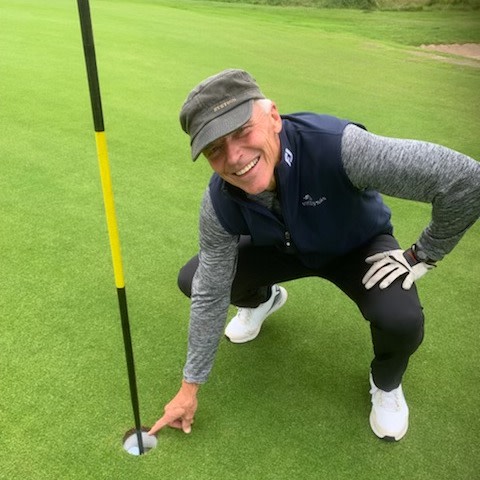 Mand med kasket og golfhandske sætter sig på hug og peger smilende på en golfbold i hullet på en golfbane.