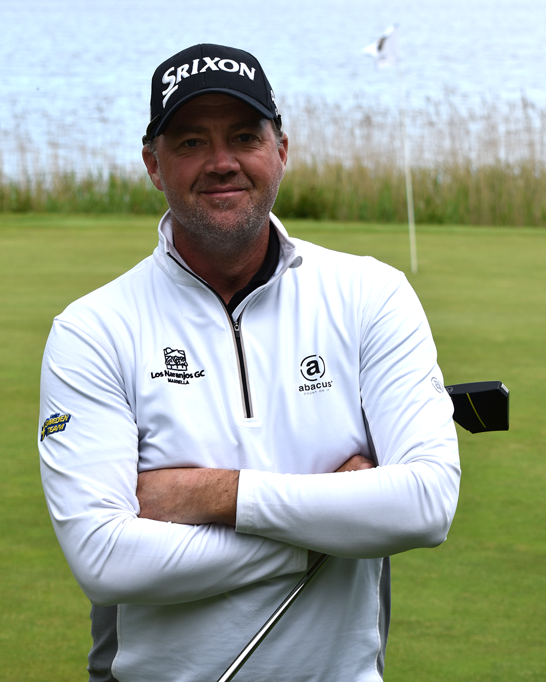 Man med golfklubba står med armarna i kors på en golfbana, bär vit tröja med logotyper och svart keps, green och vatten i bakgrunden.