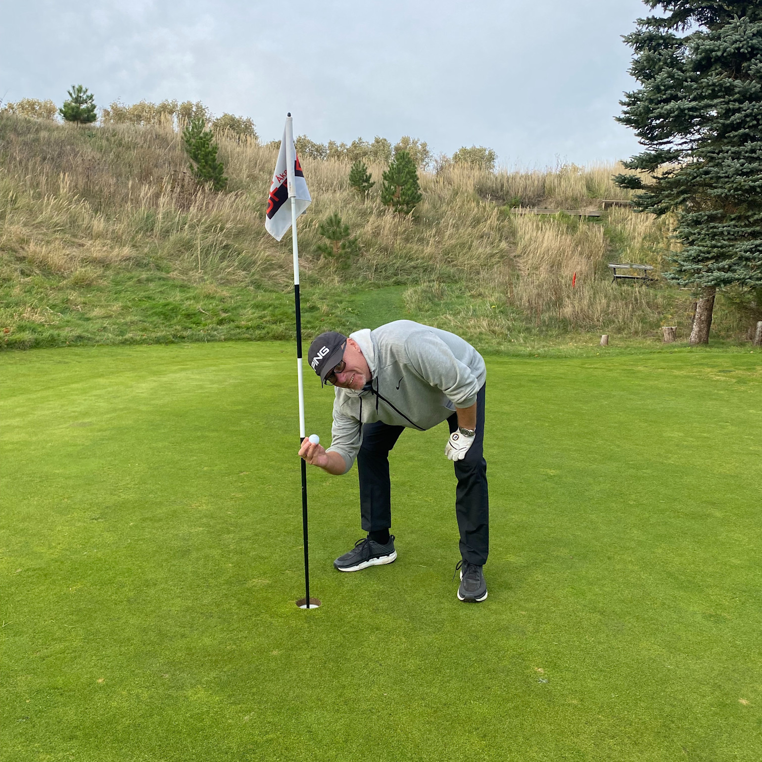 Man i golfkläder böjer sig ner vid flaggstången och plockar upp en golfboll ur hålet på en golfbana.