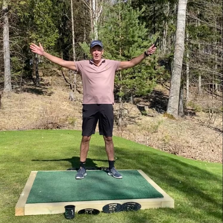 Man i sportkläder står med armarna utsträckta på en golfmatta utomhus, omgiven av träd och gräs.