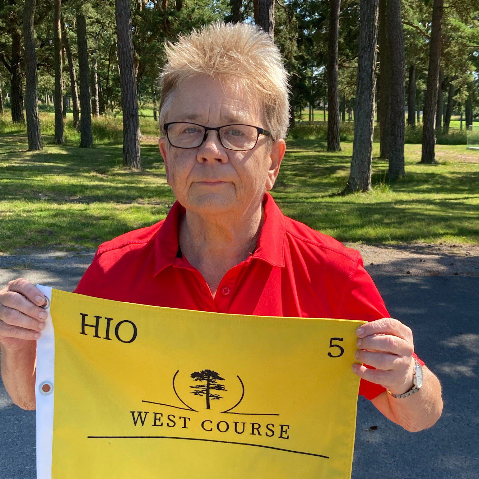 Person i rød poloshirt holder et gult flag med teksten "HIO 5 WEST COURSE" udendørs blandt træer og græs.