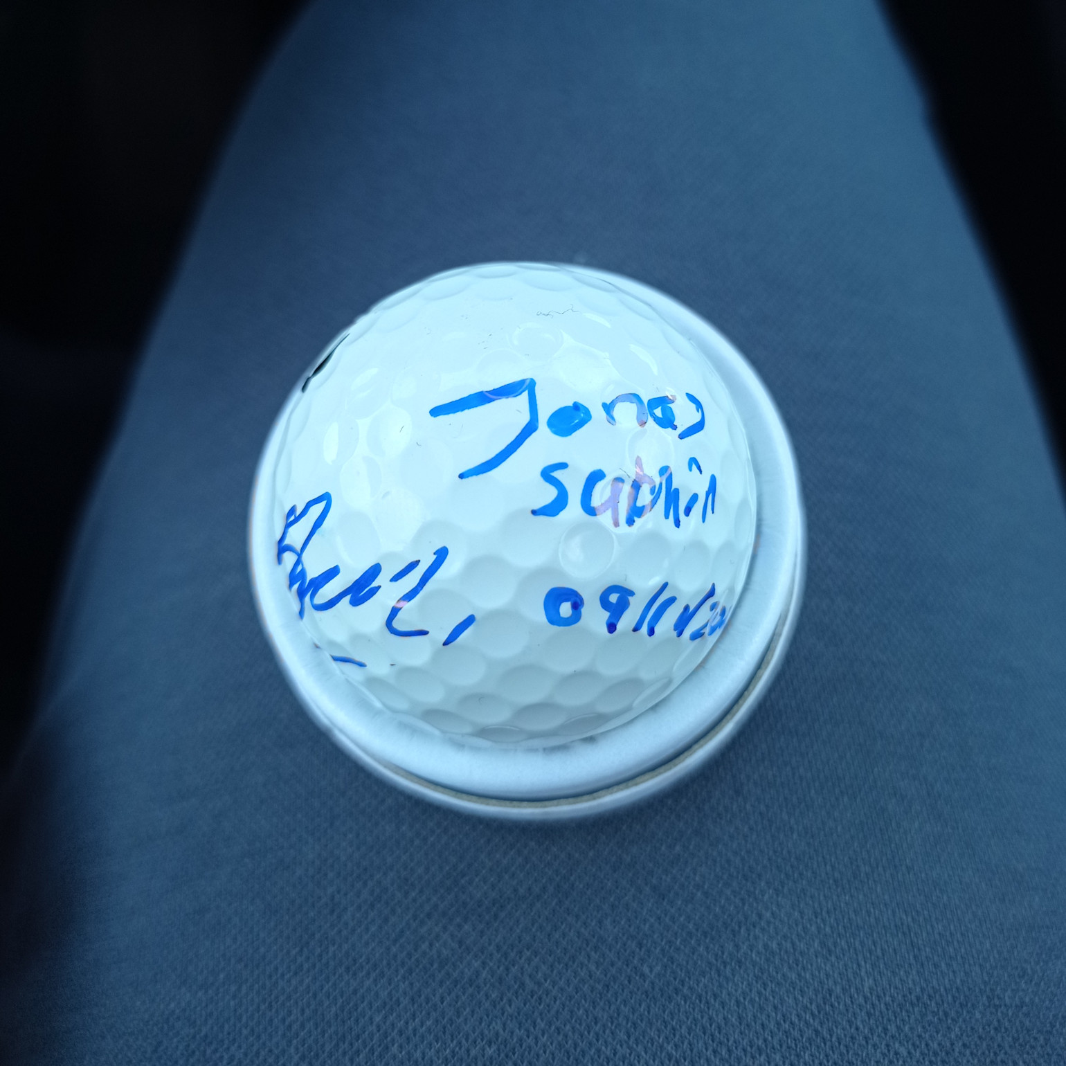 Golfbold med blå håndskrevet tekst, inklusive navnet "Jonas" og en dato, placeret på et gråt stofunderlag.