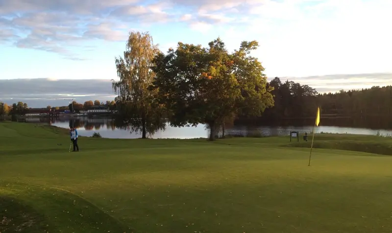En golfspelare går på en grön golfbana nära ett hål med flagga, träd och en sjö i bakgrunden.