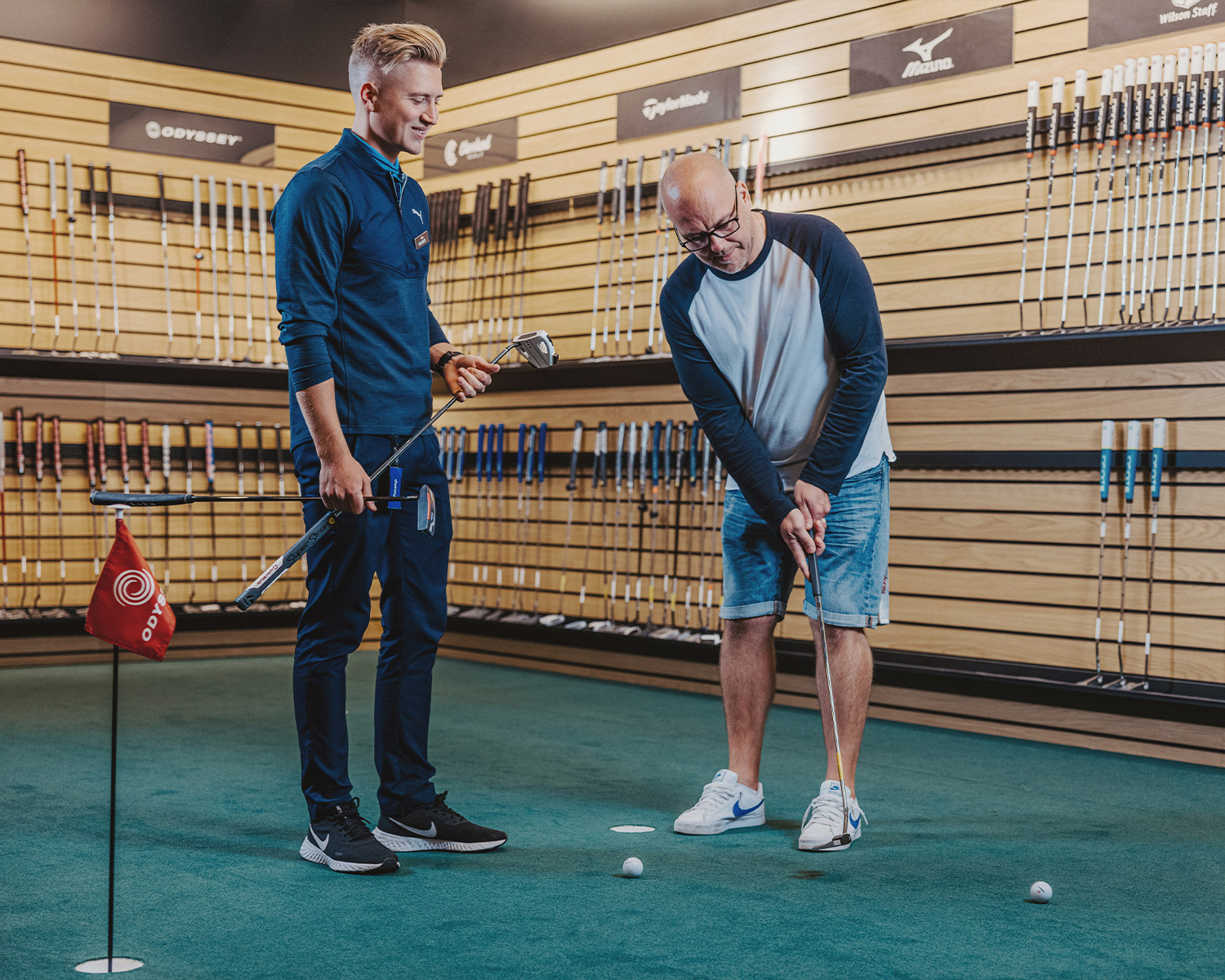 Två män provar golfklubbor inomhus; en man puttar en golfboll medan den andra håller flera klubbor och ger råd.