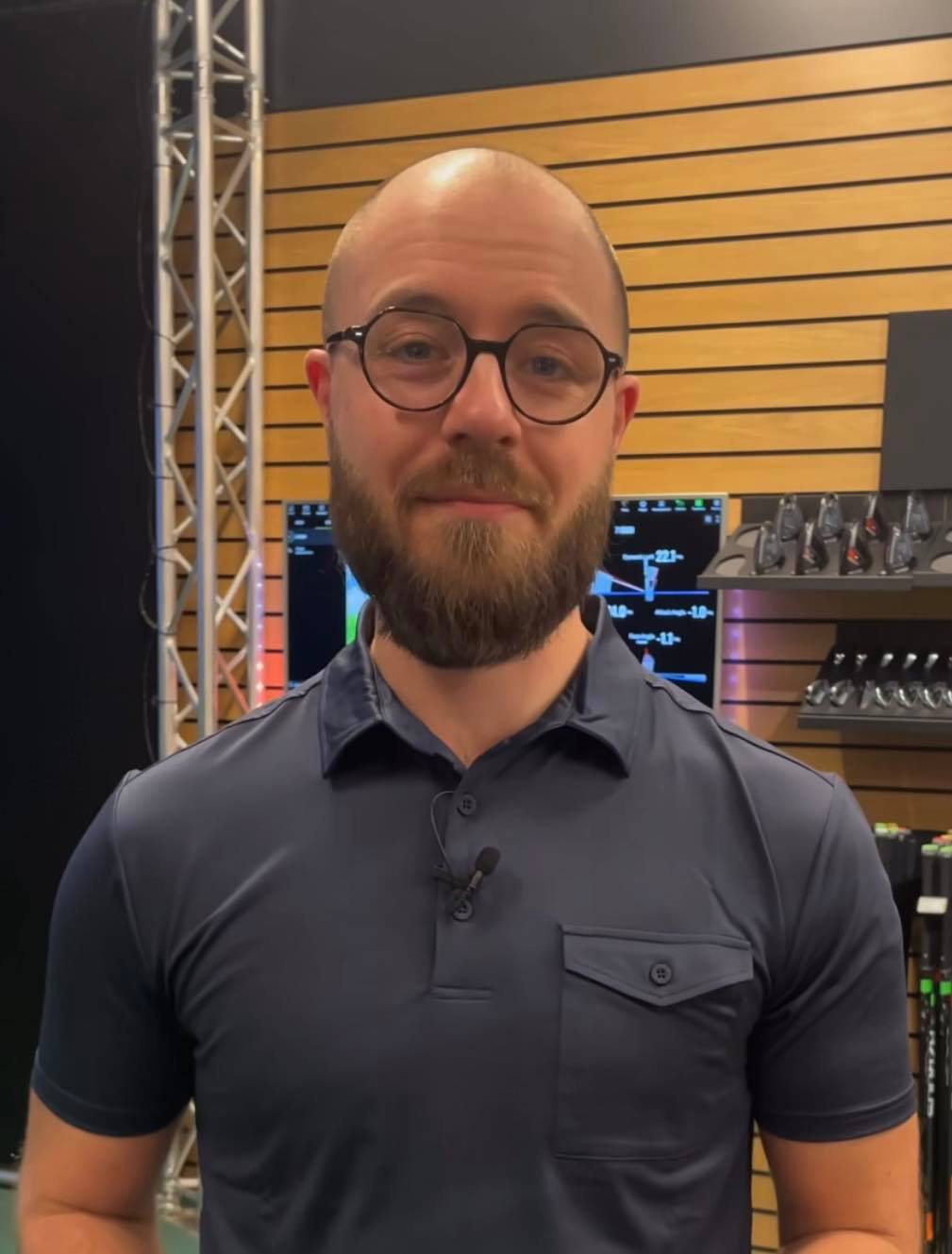 Man med glasögon och skägg står i en butiksmiljö med träpanelvägg och golfklubbor i bakgrunden.