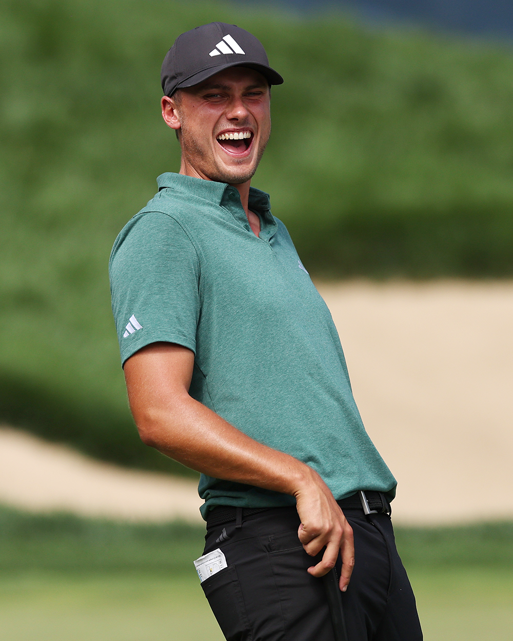 Manlig golfspelare skrattar och håller en golfklubba på en golfbana, klädd i grön pikétröja och svart keps med Adidas-logotyp.