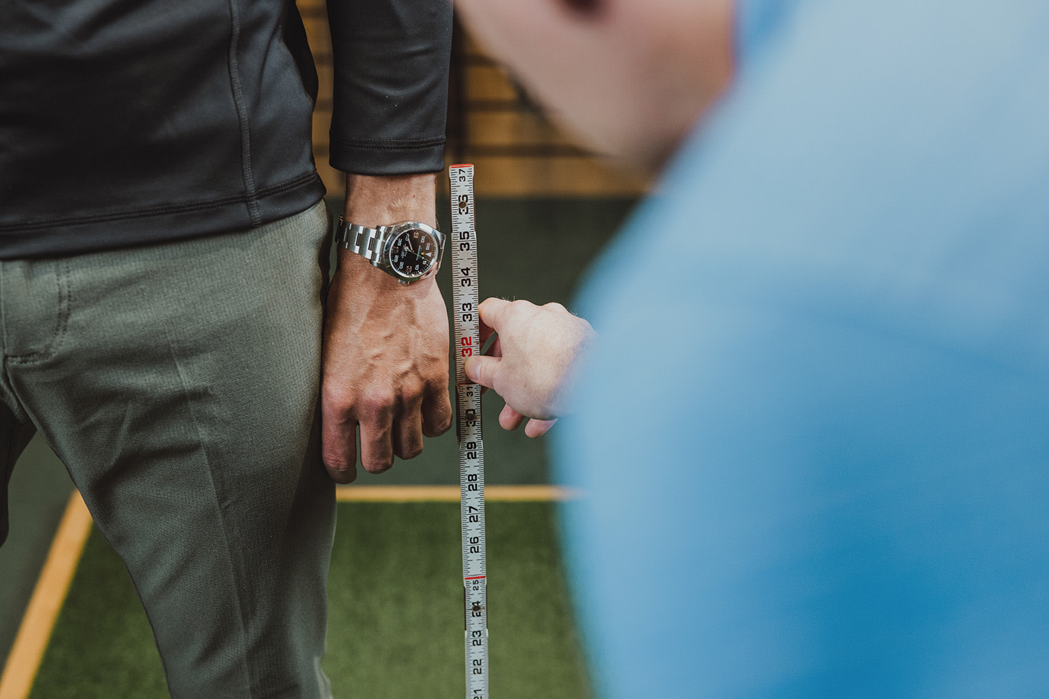 Eine Person misst den Arm einer anderen Person mit einem Maßband, wobei die eine eine silberfarbene Uhr und grüne Hosen trägt.