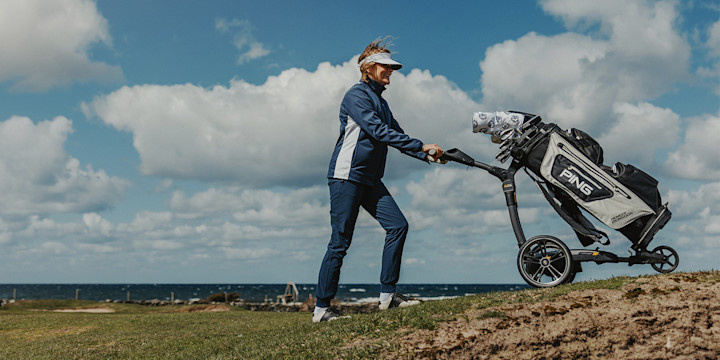 Man går på en golfbana och drar en golfvagn med golfklubbor över gräset under en klar himmel.