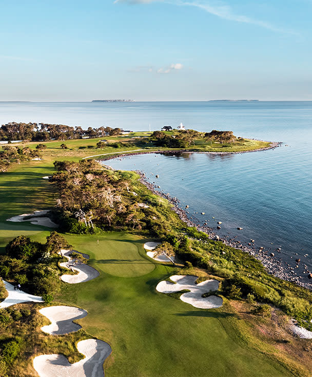 Golfbana med sandbunkrar vid kusten, grönt gräs och träd, havet breder ut sig i bakgrunden under klar himmel.