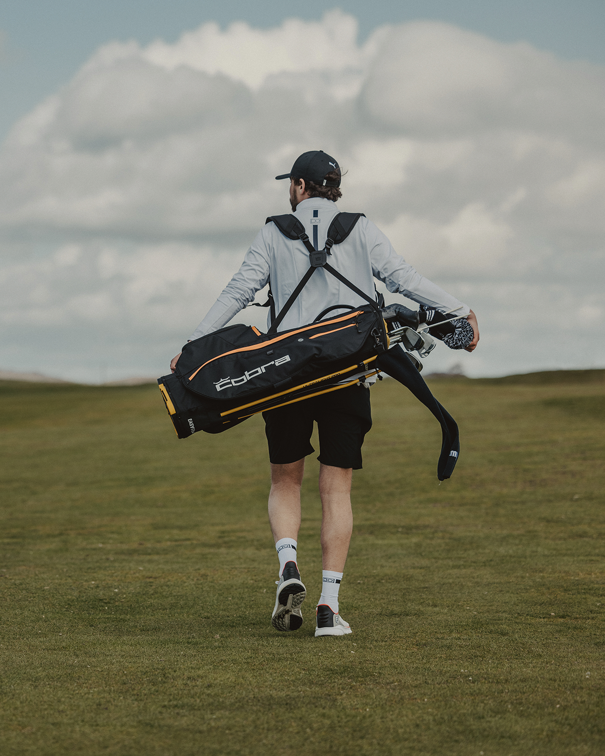 Man trägt eine Cobra-Golftasche über der Schulter und geht über einen grünen Golfplatz unter einem bewölkten Himmel.