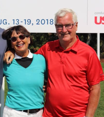 Zwei ältere Personen stehen draußen nebeneinander und lächeln, mit einem Schild im Hintergrund, das das Datum 13.–19. August 2018 zeigt.