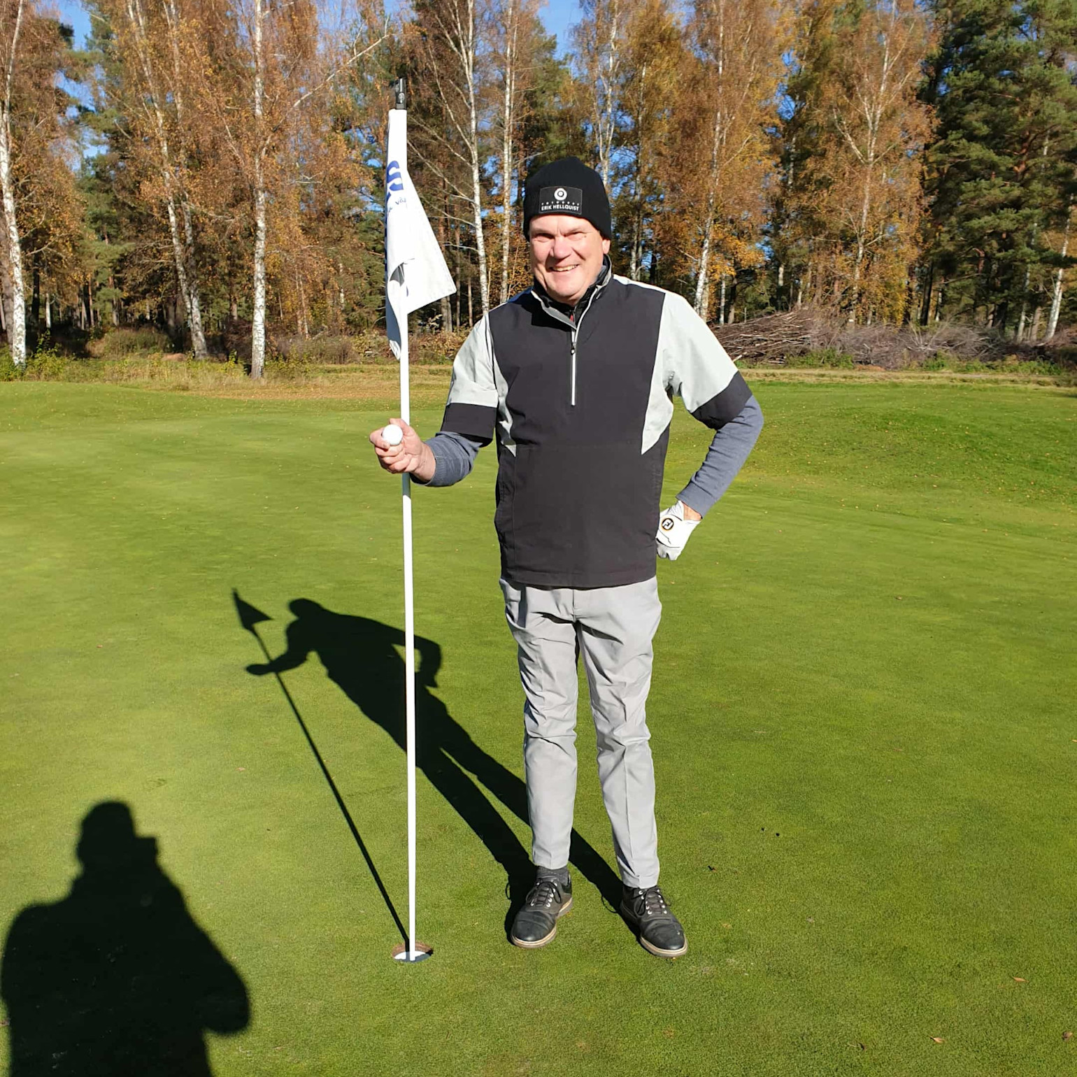 Man holder en golfball og står ved siden av en flaggstang på en golfgreen med høsttrær i bakgrunnen.