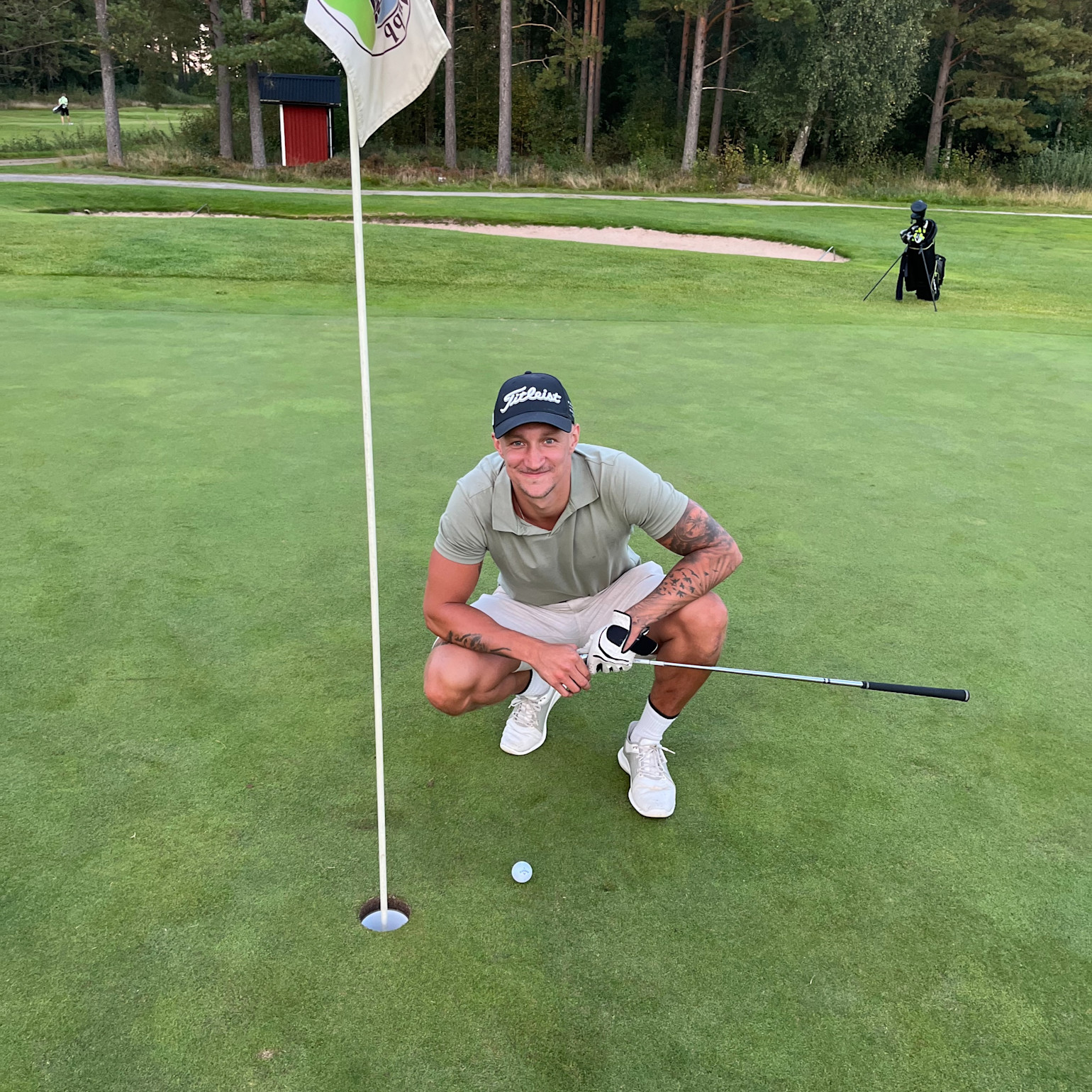 Man i golfkläder hukar sig bredvid ett golfhål med flagga, håller en klubba och ler på en grön golfbana.