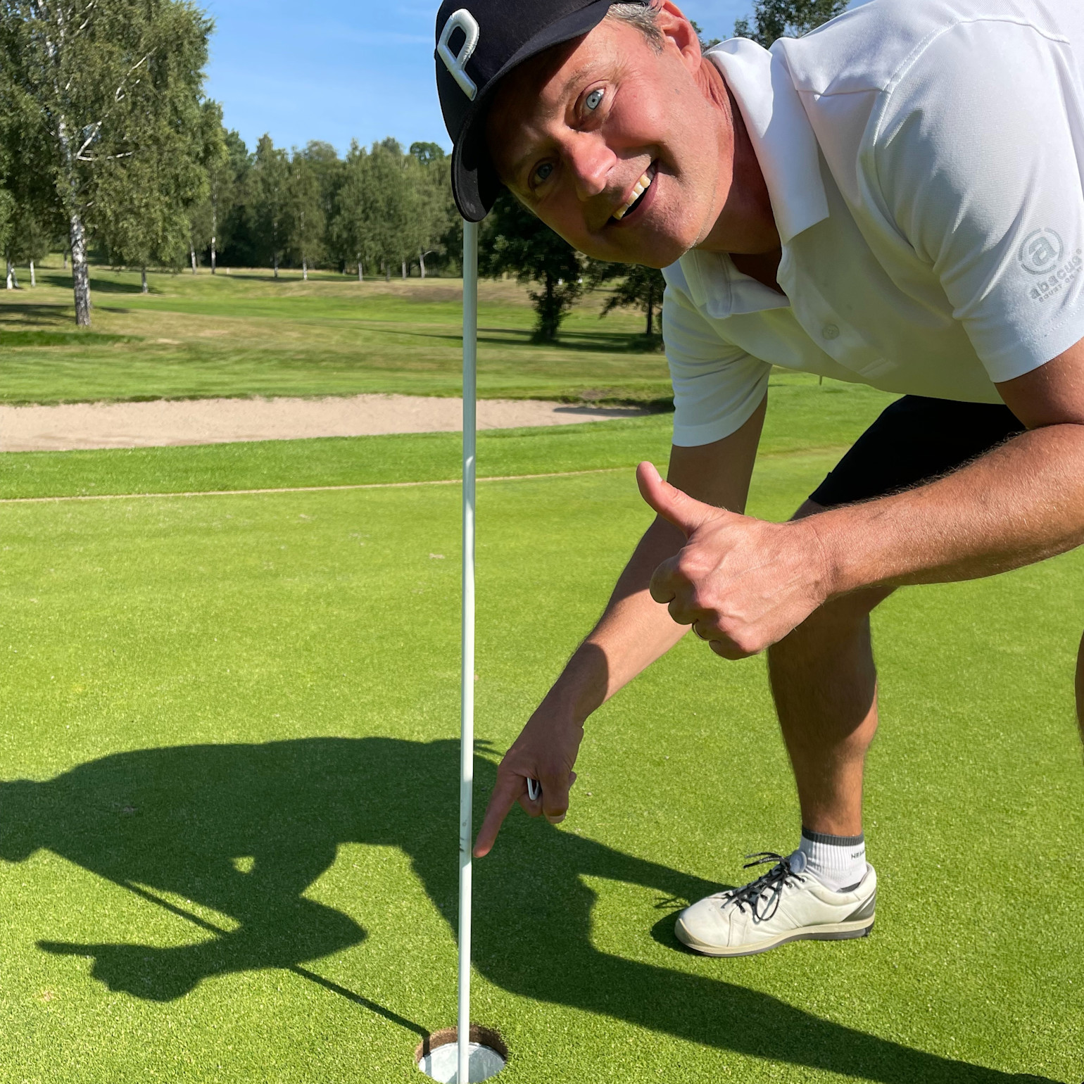 Mand iført hvid poloshirt og sort kasket peger på en golfbold i hullet og giver tommelfingeren op på en golfbane i solskinsvejr.