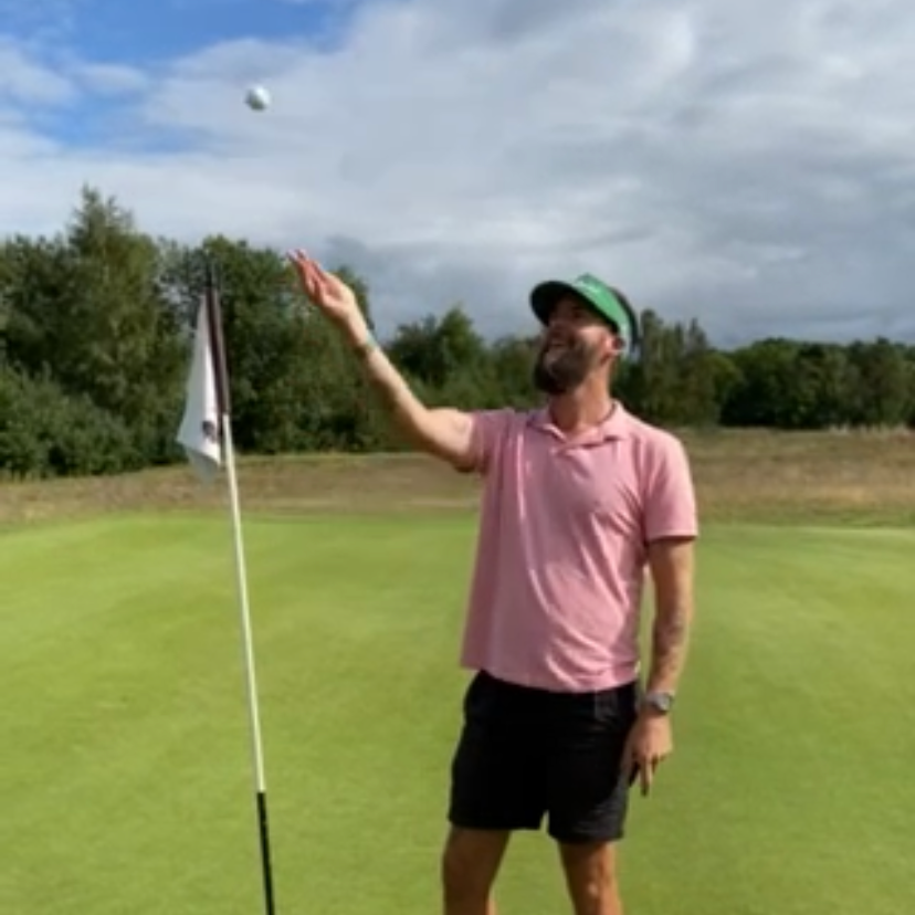 Mand i lyserød polo og sorte shorts kaster en golfbold ved siden af en flagstang på en golfbane under en overskyet himmel.