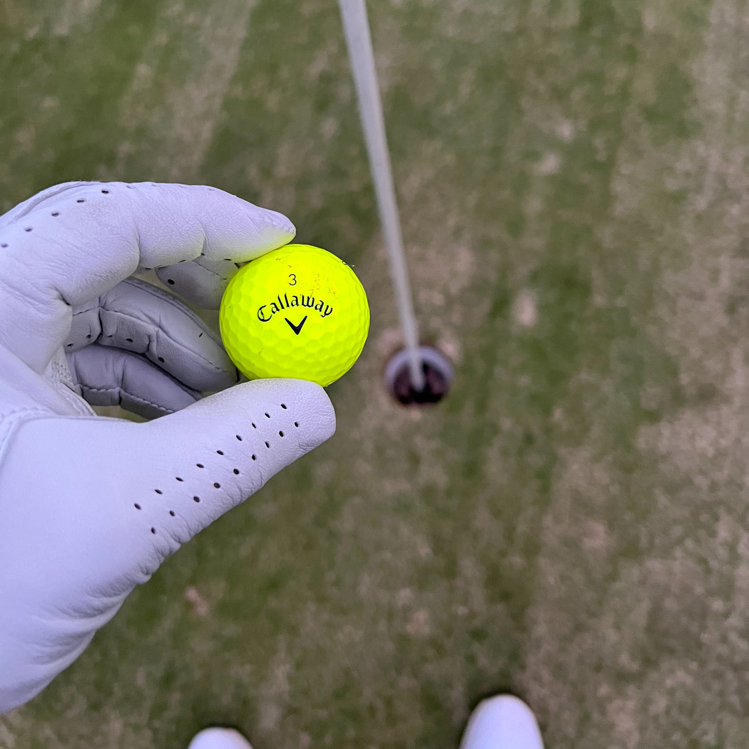 En hånd med en hvid golfhandske holder en gul Callaway-golfbold over et golfhul på en grøn puttinggreen.