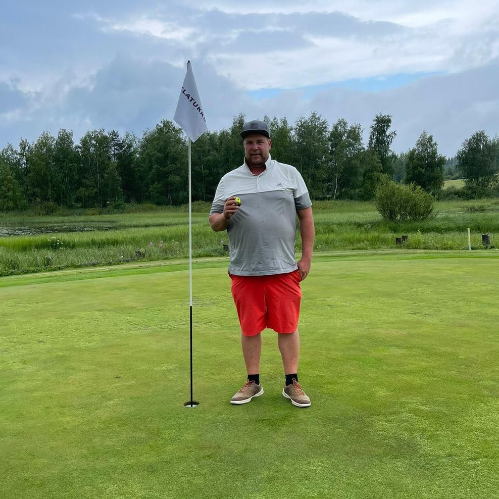 Man står på en golfgreen ved siden af et hul med flag, holder en golfbold og smiler, med træer og græs i baggrunden.