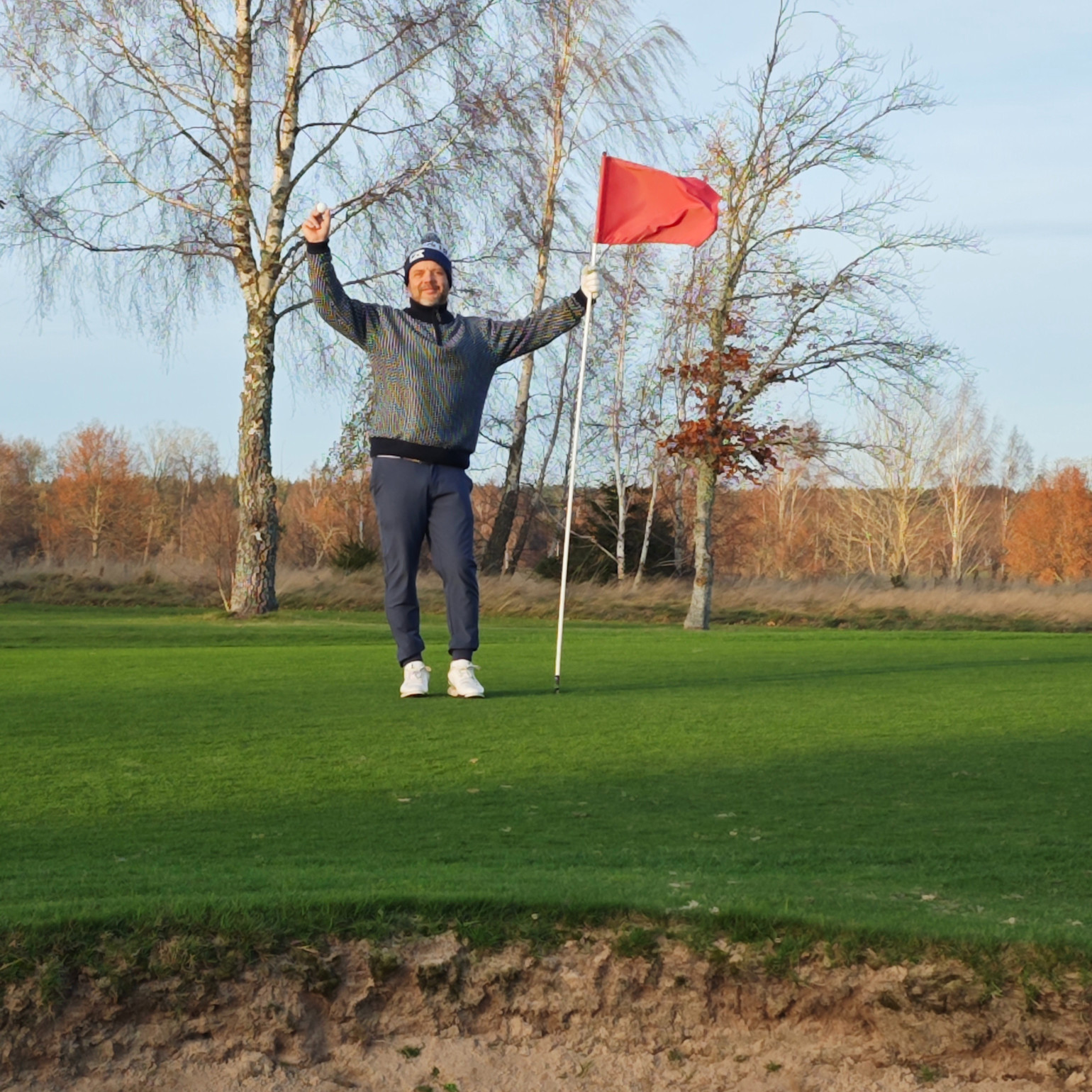 Man firar med armarna uppsträckta och håller en golfboll och flaggstång på en golfgreen, höstträd i bakgrunden.