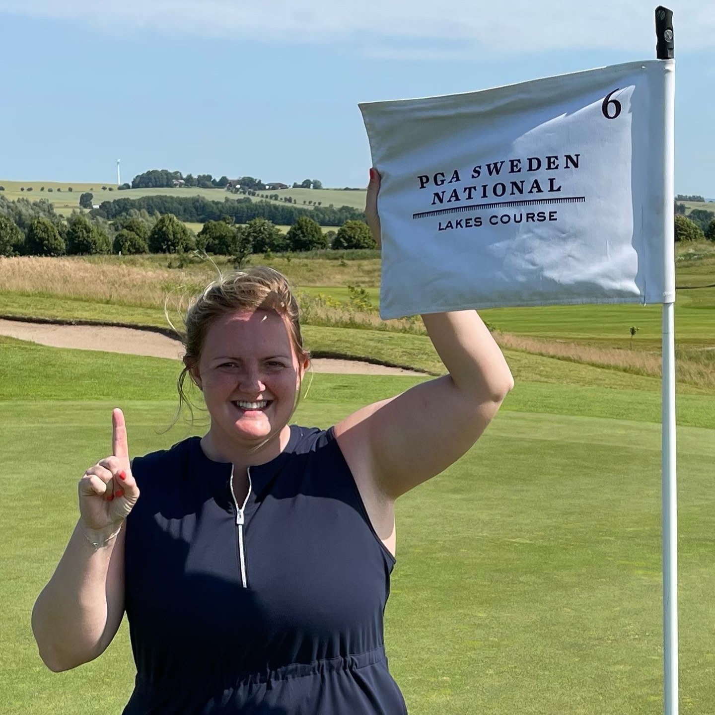 Smilende kvinde holder et flag op ved hul 6 med teksten "PGA Sweden National Lakes Course" på en solrig golfbane.