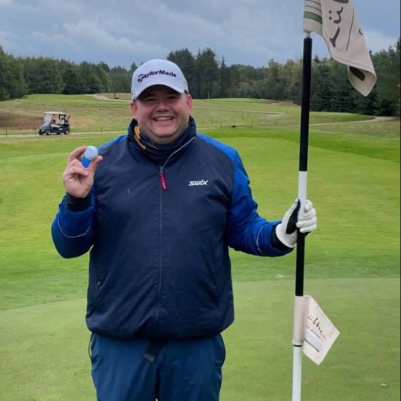 Smilende mand i golfbeklædning holder en golfbold i den ene hånd og flagstangen ved et golfhul i den anden på en golfbane.