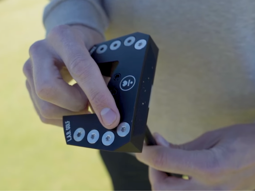 Eine Person hält einen schwarzen Golfputter-Kopf mit der Aufschrift „L.A.B. GOLF“ und mehreren silberfarbenen Gewichten am Schlägerkopf.
