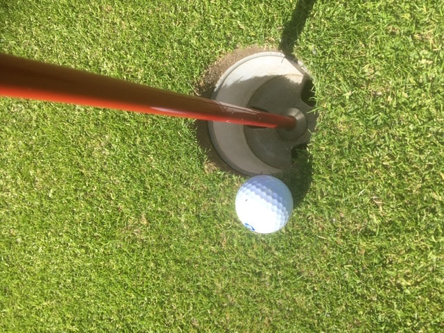 Golfbold ligger tæt på hullet på en grøn puttinggreen, med en rød flagstang placeret i koppen.