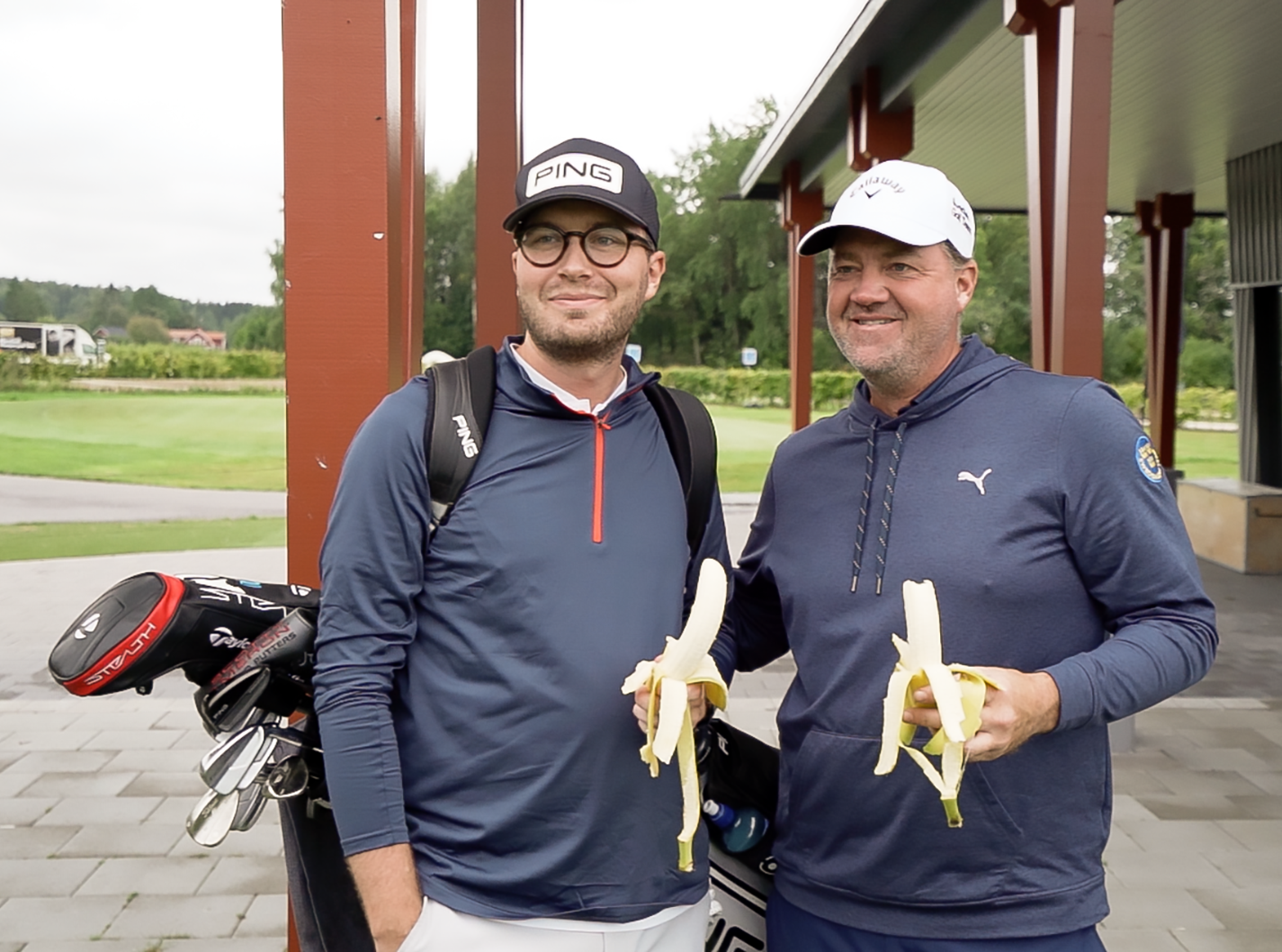 Två män i golfkläder står utomhus, håller varsin banan och ler, med golfklubbor i bakgrunden.