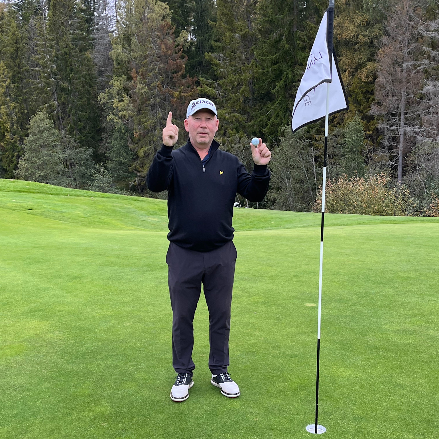 Henkilö pitää golfpalloa kädessään ja osoittaa ylöspäin lipputangon vieressä golfviheriöllä, puiden ympäröimänä.