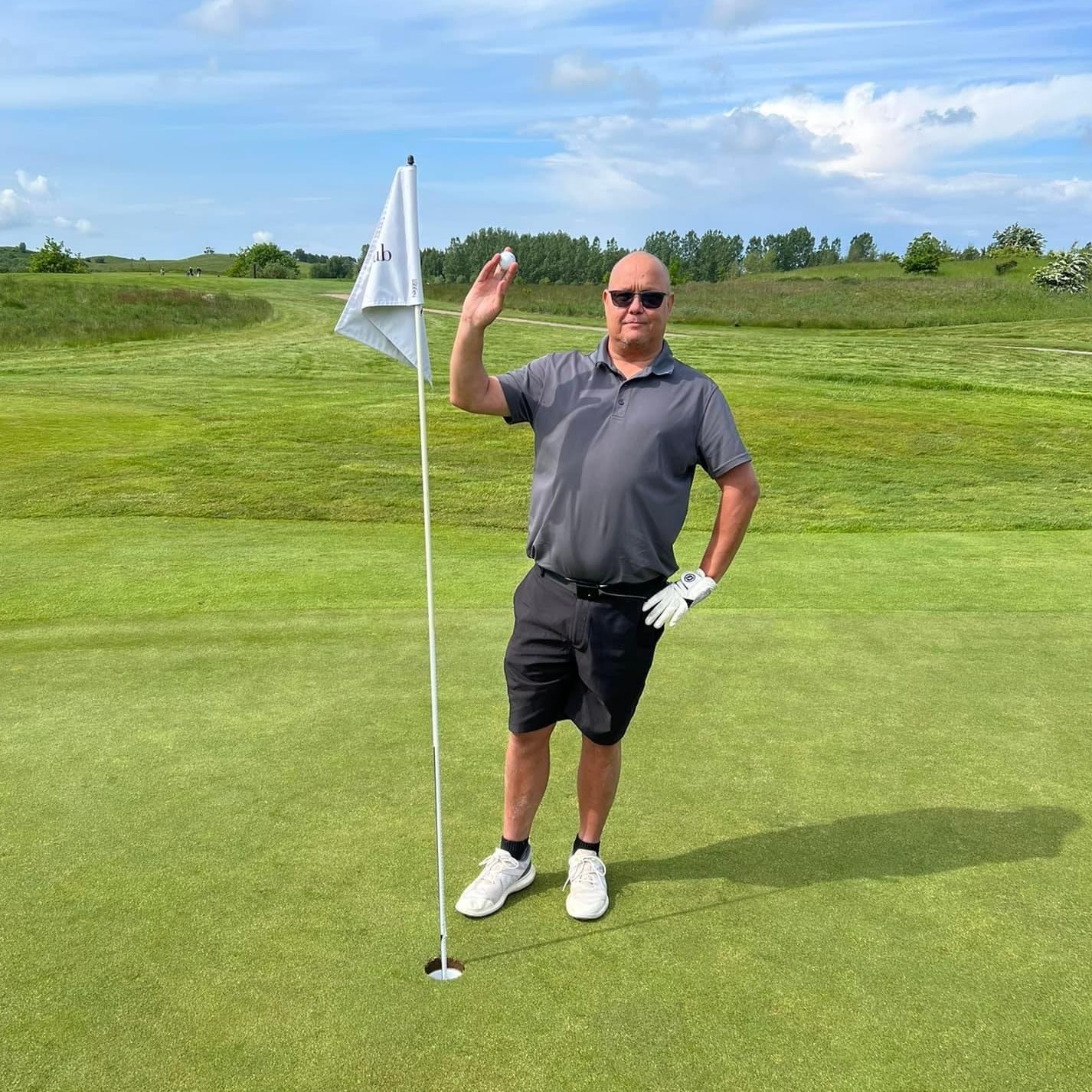 Mand på solbriller og golf­tøj står ved siden af en flagstang på en golfgreen, holder en golfbold op og smiler til kameraet.