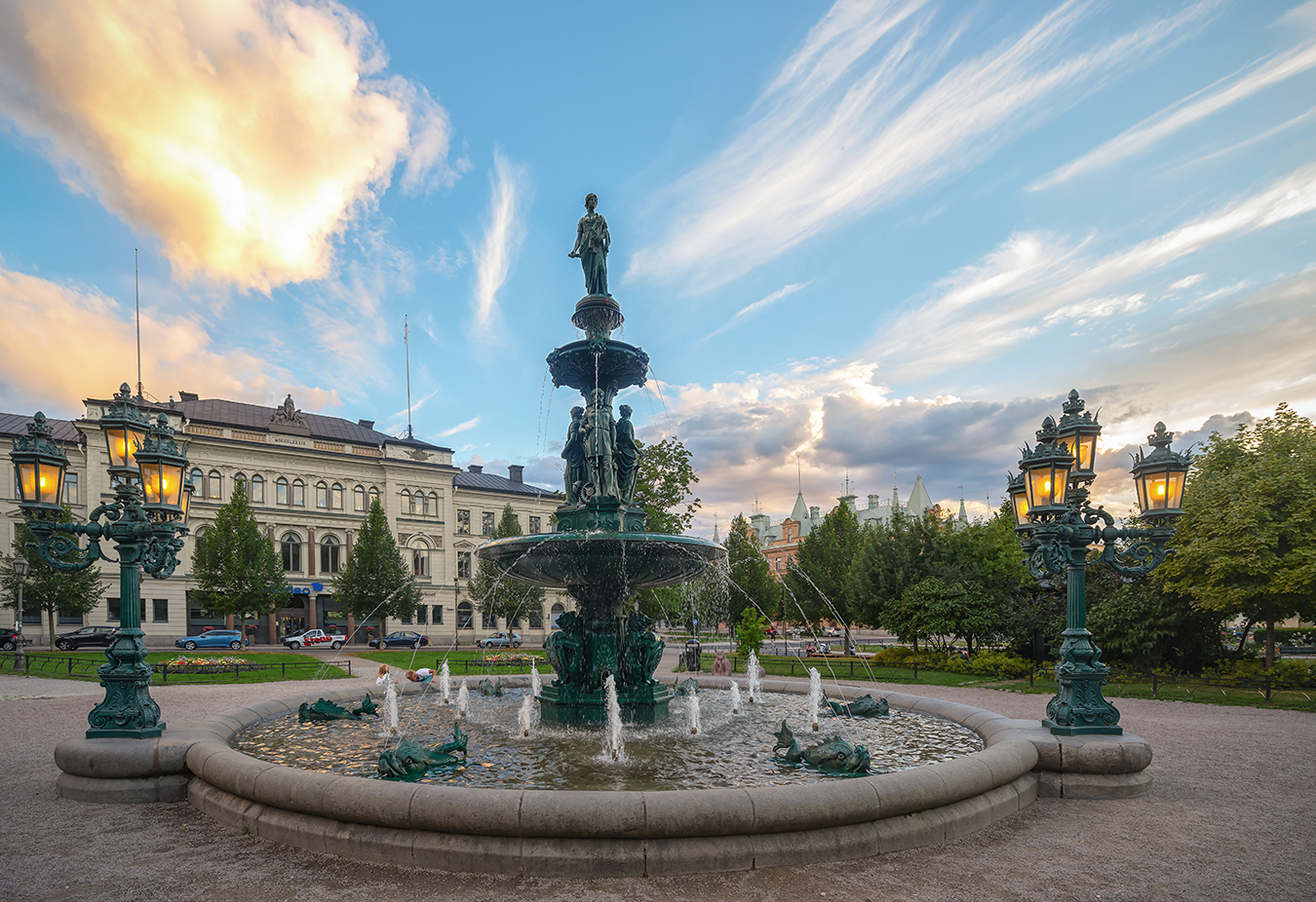 Fontän med statyer och vattenstrålar omgiven av gamla byggnader, träd och tända gatlyktor i en stadspark vid solnedgång.