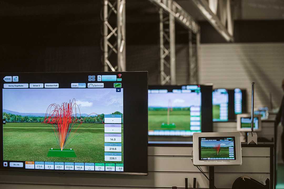Stor skärm visar golfbollarnas flygbanor och statistik i en inomhusgolfanläggning med flera liknande stationer i bakgrunden.