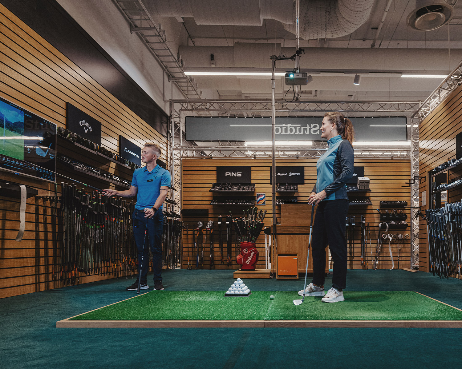Två personer står på en golfmatta i en butik, en instruerar och pekar mot en skärm medan den andra håller en golfklubba.