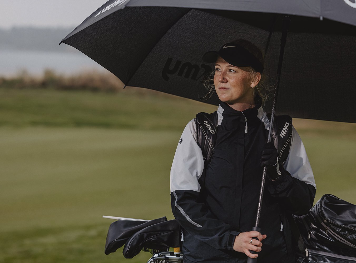 Kvinna i regnjacka står under ett stort svart paraply på en golfbana, håller i paraplyet och har golfbag på ryggen.