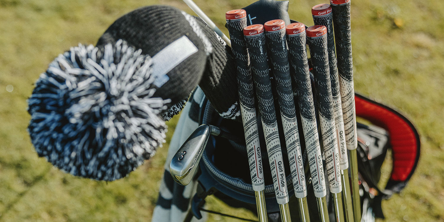 Golf clubs with Golf Pride grips stand in a golf bag next to a black and white knitted headcover on a lawn.