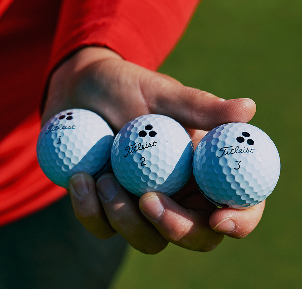 En hand håller tre Titleist-golfbollar märkta med siffrorna 1, 2 och 3 mot en grön bakgrund.