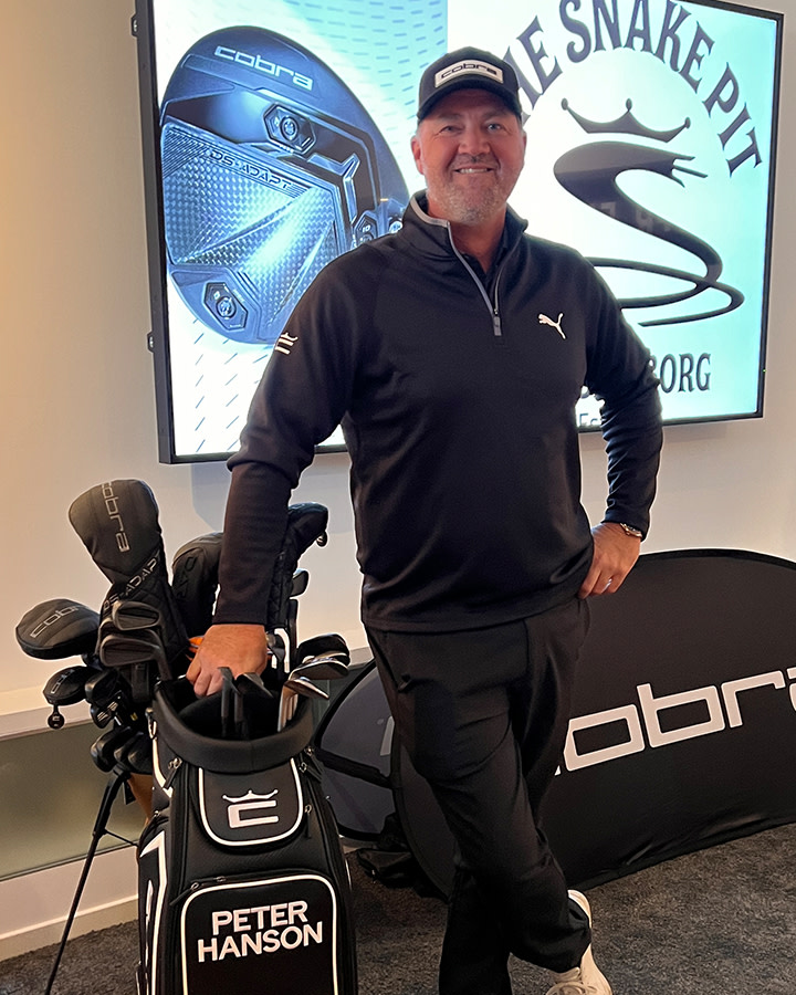 Man i svart Puma-tröja och keps står leende bredvid en golfbag märkt "Peter Hanson" med Cobra-klubbor, inomhus vid reklamskyltar.