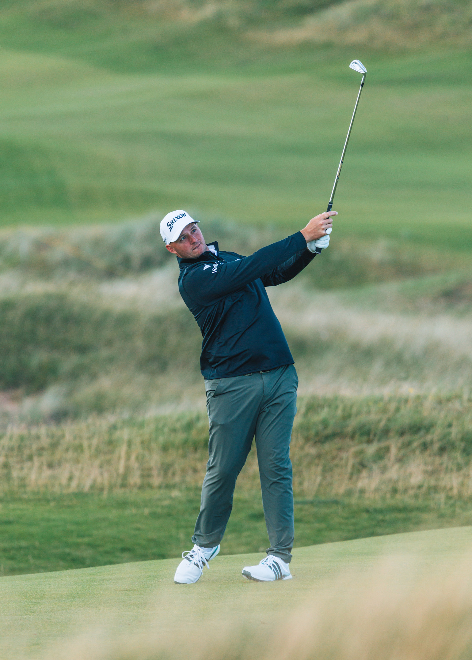 Ein Golfspieler schwingt einen Schläger auf einem grünen Golfplatz, gekleidet in dunkler Jacke, Mütze und Sportschuhen.