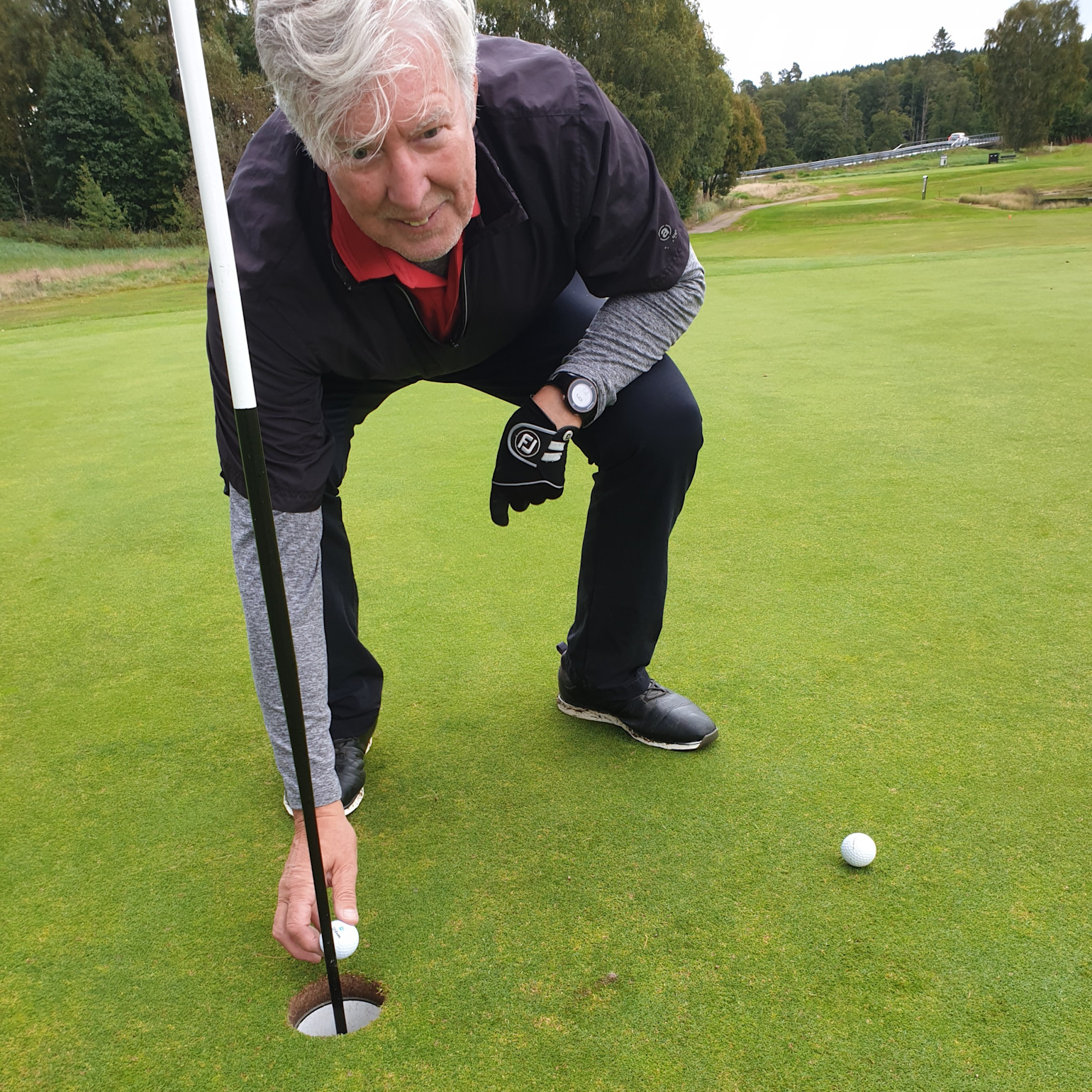 Ældre mand samler en golfbold op af hullet på en golfbane, med en anden bold ved siden af på greenen.