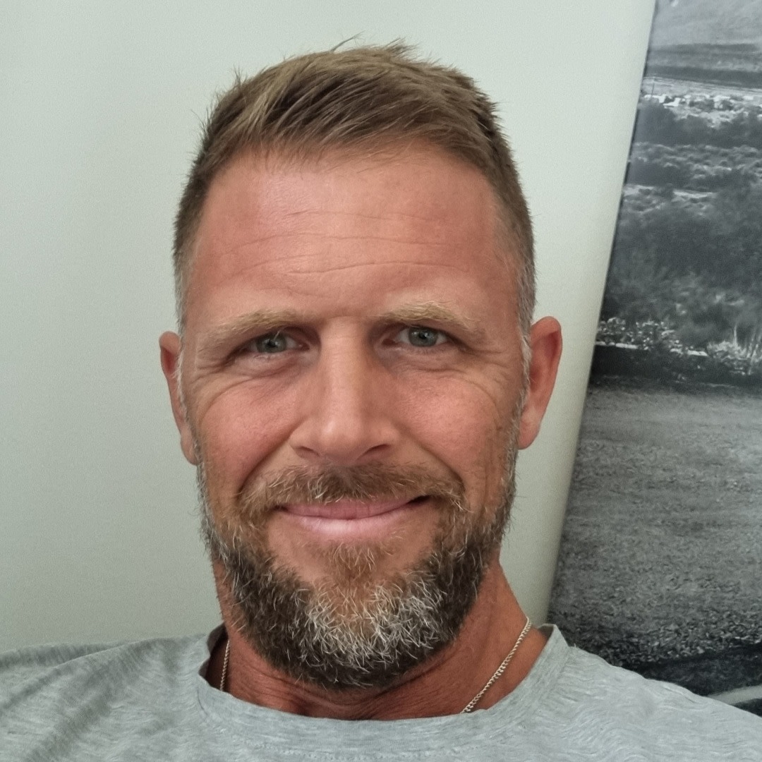 Man med kort brunt hår och skägg ler mot kameran, sitter inomhus med en tavla i bakgrunden.