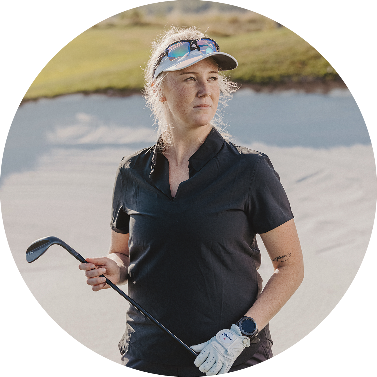 Kvinna i svart golftröja och keps håller en golfklubba och står i en sandbunker på en golfbana.