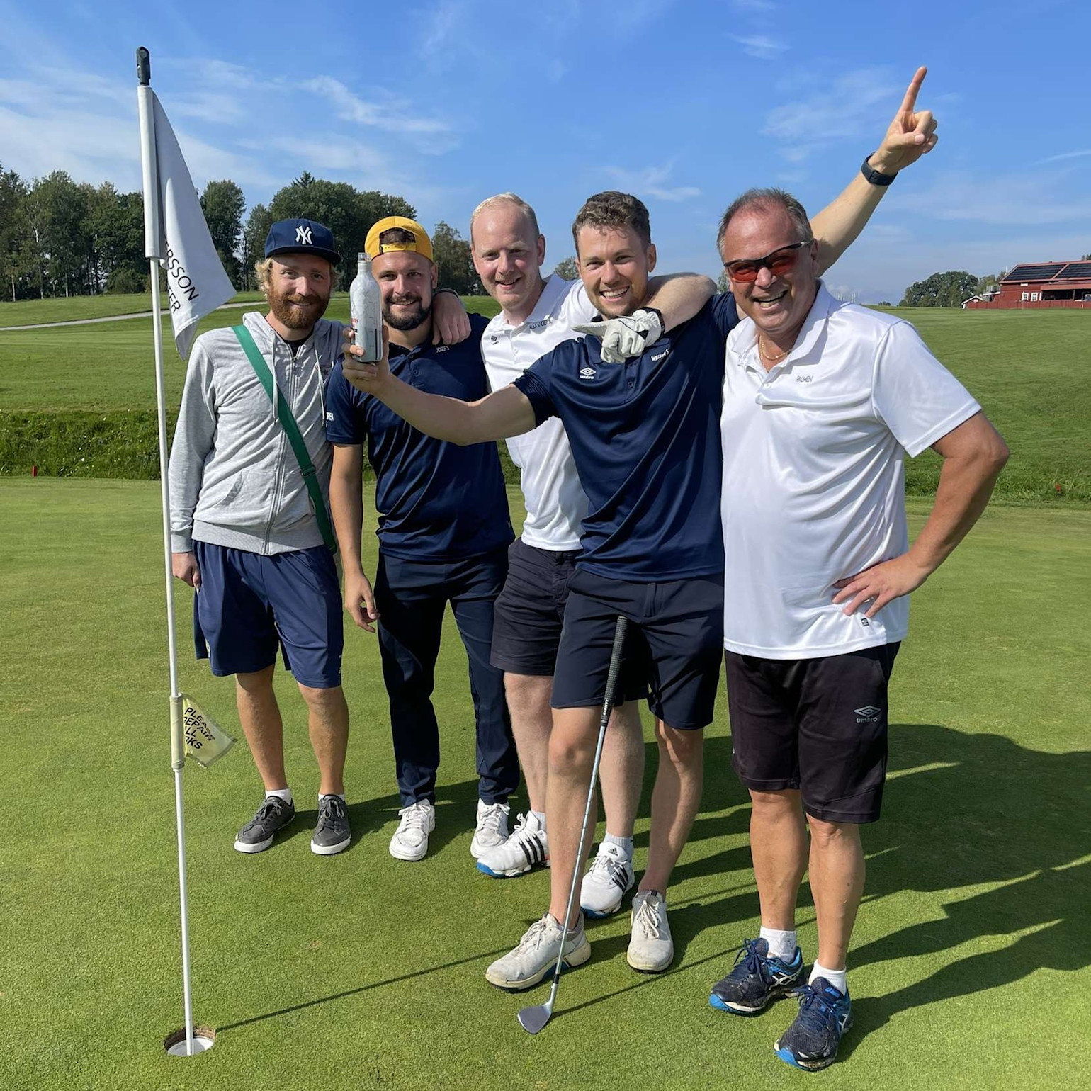 Fem glada män poserar tillsammans på en golfbana vid hålet, en håller en golfklubba och visar en burk mot kameran.