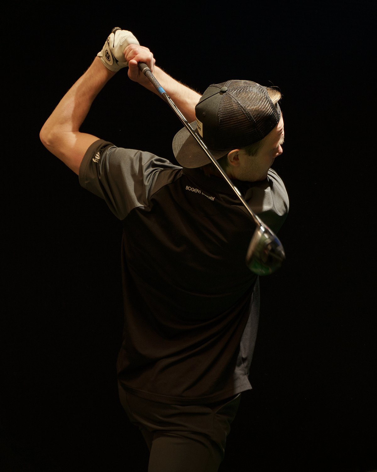 En person i keps och golfhandske svingar en golfklubba bakåt, mot mörk bakgrund.