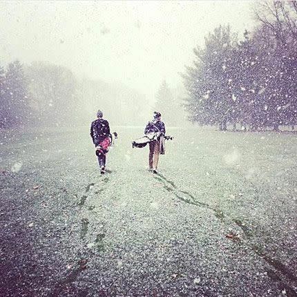 Två personer går på en snötäckt golfbana och bär golfbagar medan stora snöflingor faller omkring dem.