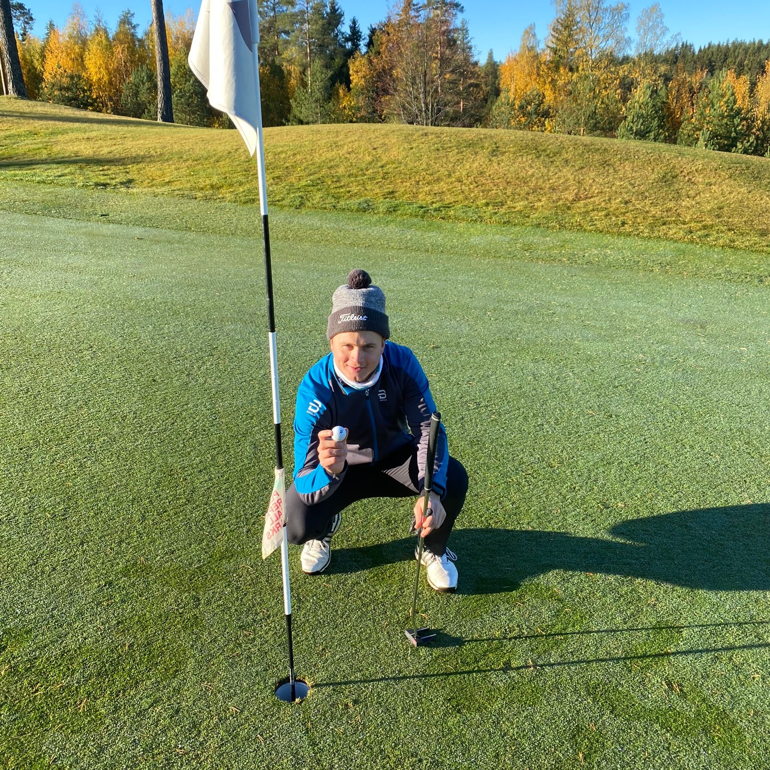 Person i blå jakke og hue sidder på hug ved et golfhul, holder en golfbold og en putter på en solrig golfbane med efterårstræer i baggrunden.